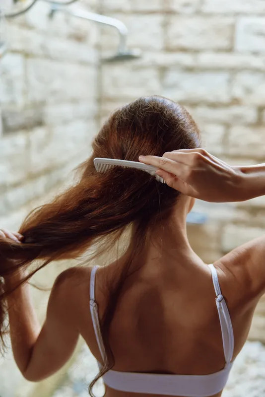 Woman combing long wet hair in bright bathroom with stone wall background, natural beauty care routine, personal grooming moment, close-up rear view.