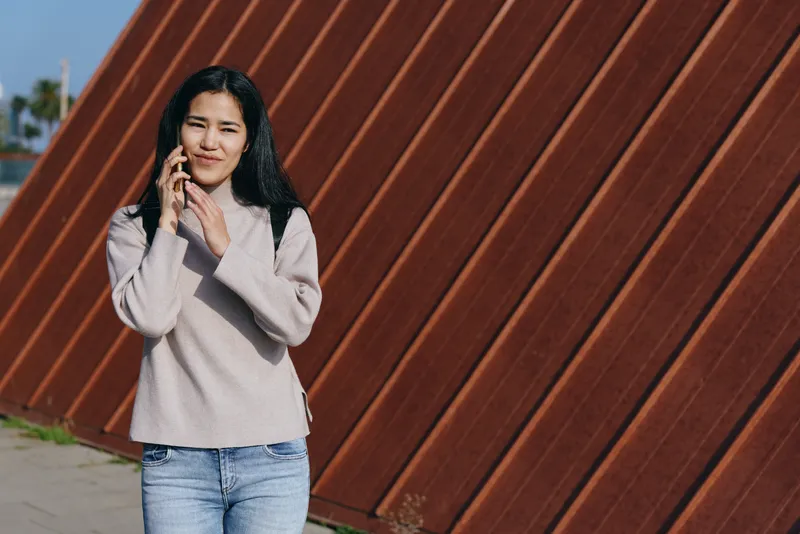 Casual scene of a woman outdoors beside a rusted wall, talking on a mobile phone, wearing a light sweater and jeans, highlighting everyday communication, relaxed mood, street style.