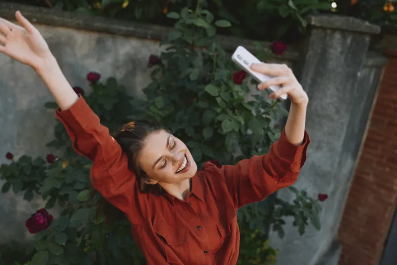 Young woman in a red blouse takes a joyful selfie outdoors among blooming roses, capturing a candid lifestyle moment with natural light. Bright mood brightens portrait