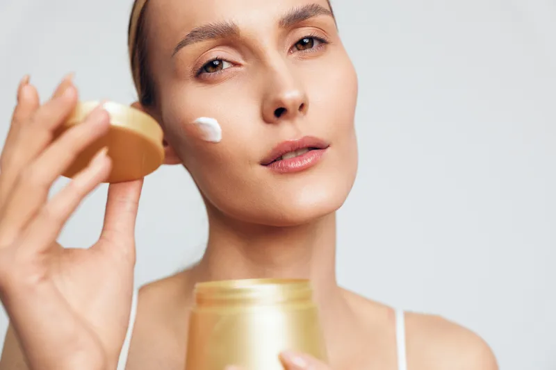 Closeup of woman applying face cream with fingers holding open jar skincare treatment natural beauty routine isolated on white background with soft light and calm expression