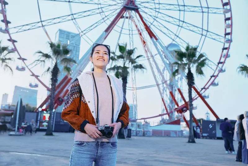 Woman outdoors with a camera at an amusement park, ferris wheel and palm trees in the background, capturing a vibrant travel photography moment amid city lights
