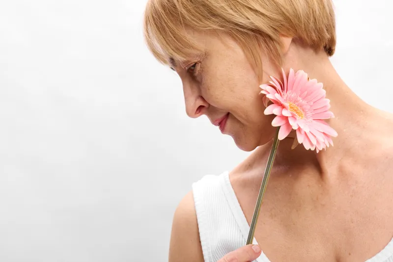 Woman closeup holding pink flower to neck, tenderness and beauty, calm mature female portrait on white background, isolated with soft light