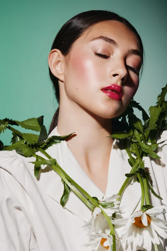 Portrait of young woman with glossy pink lipstick and flawless skin surrounded by white flowers on green background. Creative beauty and fashion concept with natural vibe and soft light.