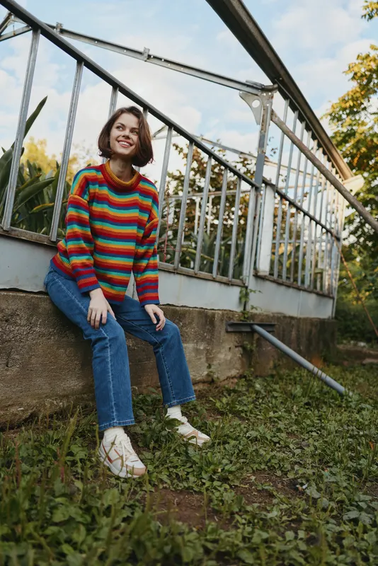 woman, colorful sweater, jeans, sitting, smiling, short hair, outdoor: young woman in her 20s wearing a rainbow striped sweater and ripped jeans, sitting on a concrete ledge by a metal railing,