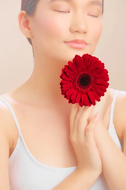 Asian woman with a serene expression holds a bold red flower near her face, exuding calm beauty and gentle grace in a soft studio ambiance, inviting tranquility and poise.