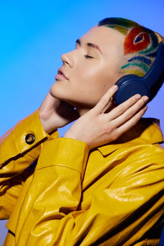 Young woman with colorful hair design wearing yellow jacket and headphones, enjoying music with closed eyes, vibrant modern style, studio blue background.