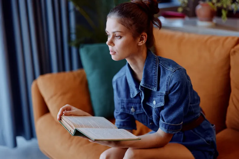 Женщина в джинсовой одежде читает книгу на диване в уютной гостиной