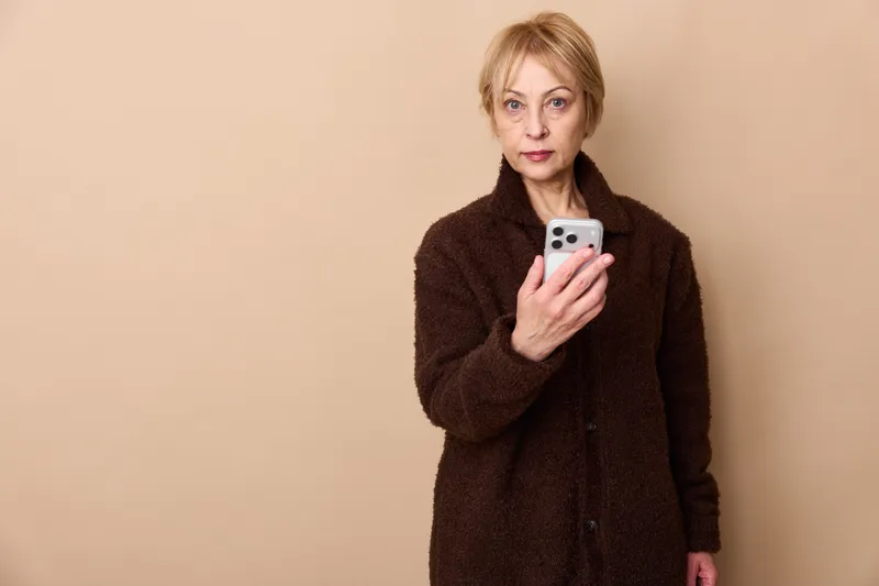 Woman holding smartphone, studio portrait, technology and communication, online messaging, texting, elderly mature blond woman, neutral beige background, expression