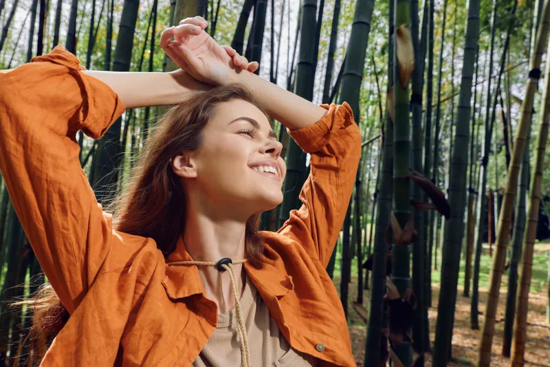 Woman in a bamboo forest smiles with eyes closed, sunlight on her face. Nature portrait shows relaxation and happiness outdoors, peaceful moment in green woodland.