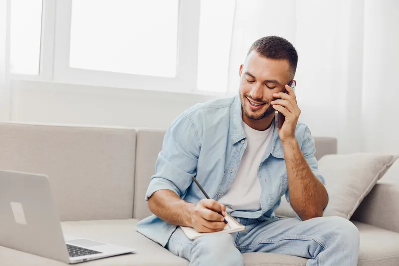 Smiling man engaged in a phone conversation while taking notes at home. Bright, modern interior emphasizes productivity and comfort in a cozy workspace.