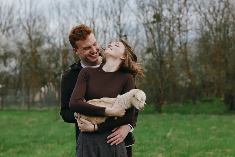Cheerful couple outdoors with a small goat kid, embracing and laughing, sunlit meadow setting, warm atmosphere, candid affection, natural colors, rustic backdrop, casual lifestyle moment