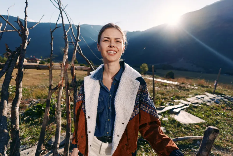Woman smile outdoors in mountains, rural setting by wooden fence with sunlight backlight, portrait of a casually dressed woman in jacket and denim enjoying scenic countryside.