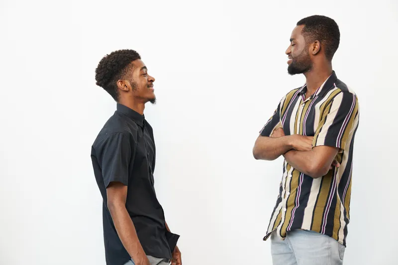 Two black men engaged in conversation, sharing smiles and laughter. The photo captures a moment of friendship against a clean white background.