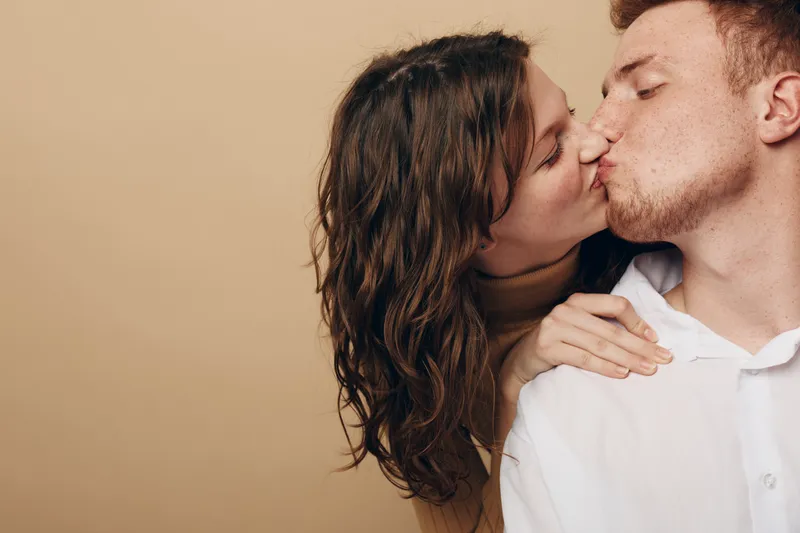 Young couple kissing tenderly with closed eyes, woman holding man’s shoulder. Intimate moment captured on beige background. Concept of love, affection, and romantic relationship.