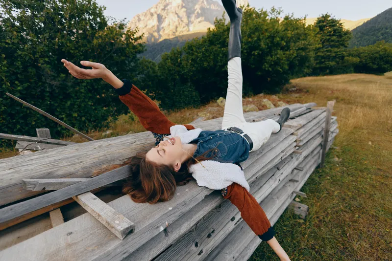 Woman relaxing on wooden fence in meadow near mountains, casual autumn outfit and boots, lying back with arms outstretched, outdoor leisure and joyful nature escape.