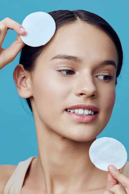 Young woman with a natural look holding cotton pads, radiating confidence and freshness against a bright blue background, perfect for skincare and beauty concepts