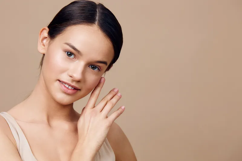 Young woman with smooth skin and natural makeup touching her cheek, expressing confidence and beauty, on a warm beige background