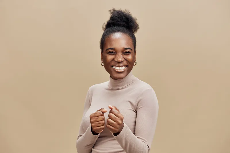 Happy young woman with curly hair smiling joyfully against a beige background, showcasing a cheerful emotion in a stylish turtleneck