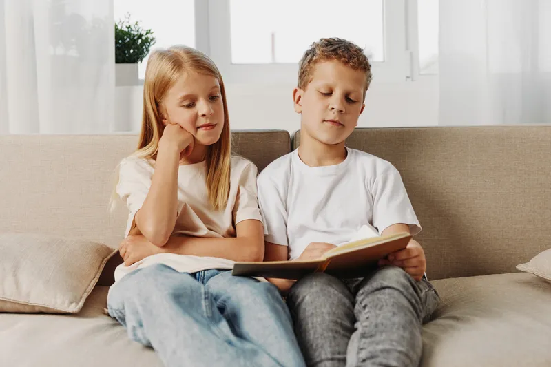 Two children sit on a beige couch reading a book together in a bright room with natural light. The boy and girl focus on the story, sharing a quiet, peaceful moment of learning.