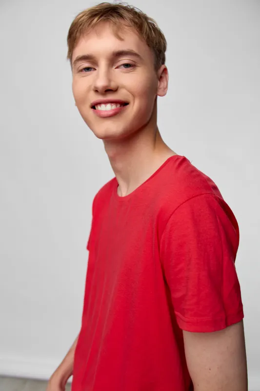 A smiling young man in a red shirt stands in a bright studio, exuding a casual and confident vibe. Clean lighting highlights his friendly expression, ideal for lifestyle, fashion and contemporary