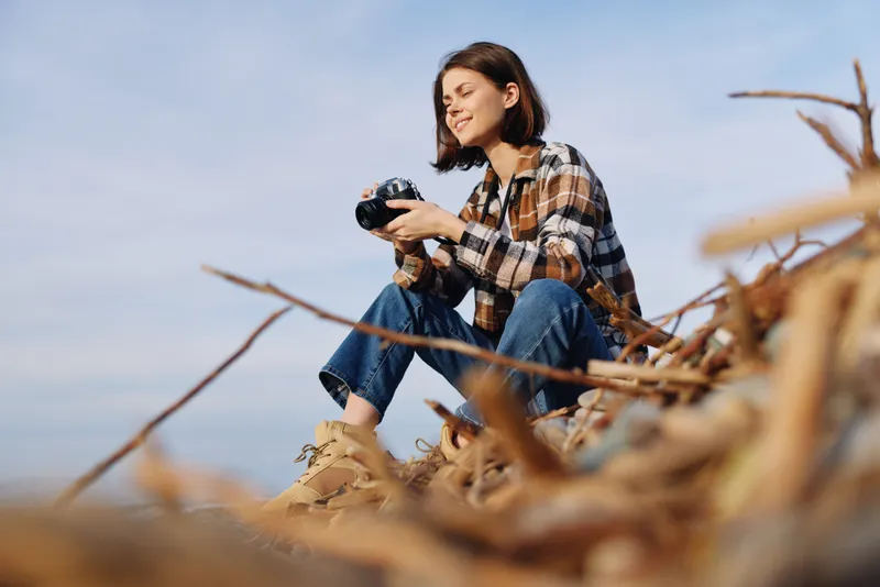 Young woman sits on sandy dunes with a camera, enjoying an outdoor photography moment, dressed in a plaid shirt and jeans, capturing coastal scenery and the calm sky during a sunny day.