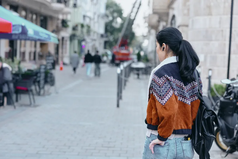 Street scene with a woman seen from behind, wearing a patterned orange jacket, jeans and a backpack, strolling along a quiet city sidewalk on a cool day, capturing urban style and casual confidence