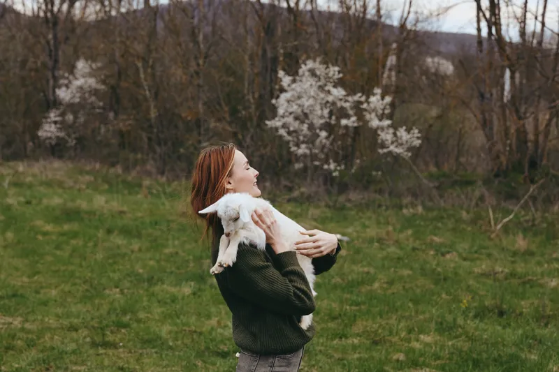 A woman outdoors in a green field holds a small goat kid close, smiling softly. Warm light and blooming trees frame this tender moment of countryside bonding and gentle animal care.
