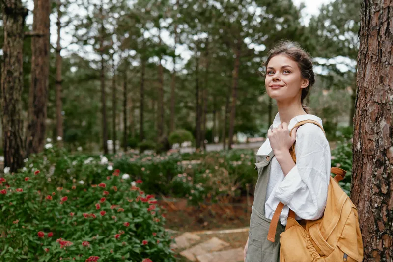 Young woman in casual outfit with a backpack, smiling outdoors surrounded by greenery and flowers, capturing a serene moment in nature