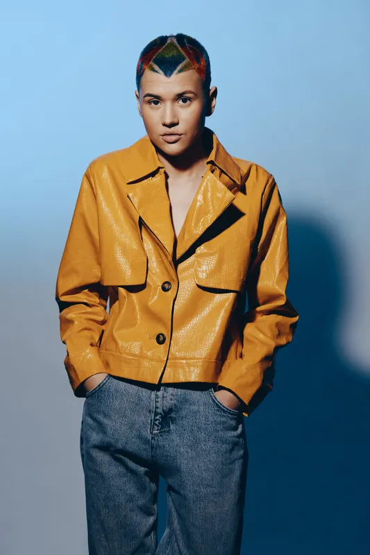 young woman with colorful short hair wearing mustard yellow jacket and jeans, standing against gradient blue background with a confident pose and serious expression
