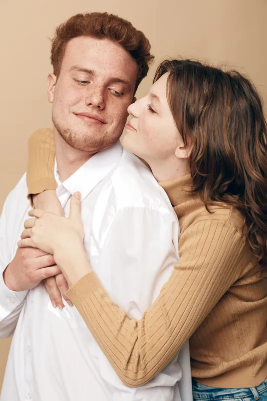 Young couple embraces tenderly in casual clothes against beige background. Woman hugs man from behind showing affection and warmth. Concept of love, relationship, intimacy, and connection.