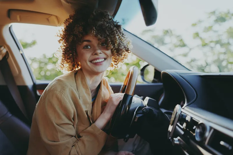 Woman in car, lifestyle moment with warm sunlight, relaxed smile, hands on steering wheel, casual fashion, natural colors, cozy interior, candid expression, daytime drive