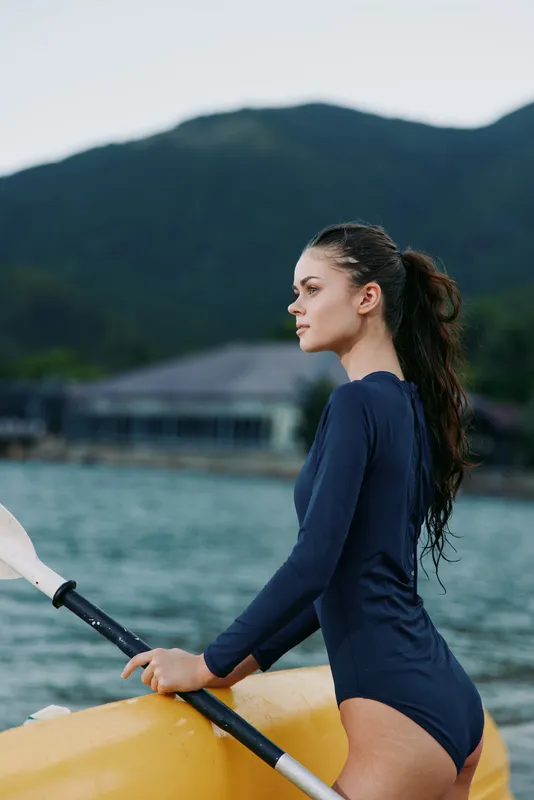 Young woman in a navy one piece stands beside a bright yellow kayak on calm water, gripping a paddle as distant mountains rise, creating a serene lake scene.