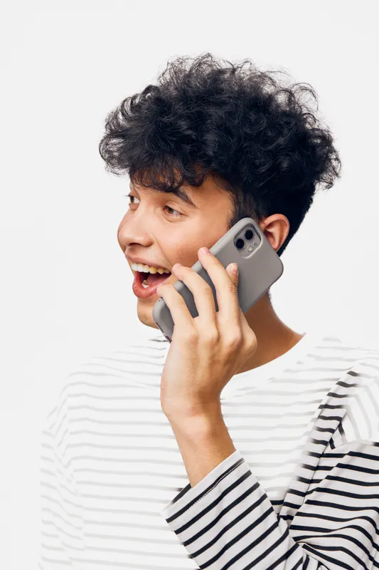 Young man talking on smartphone with joyful expression, curly hair, casual striped shirt against white background, communication and technology concept.