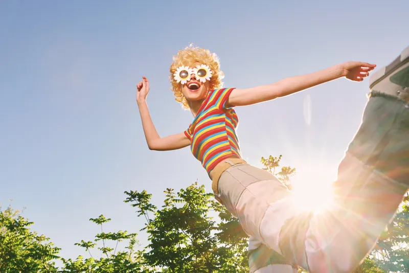 Joyful woman with striped top and flower sunglasses jumping outdoors against a clear blue sky, sunlight flare, capturing a playful retro lifestyle and vibrant summer mood with candid energy