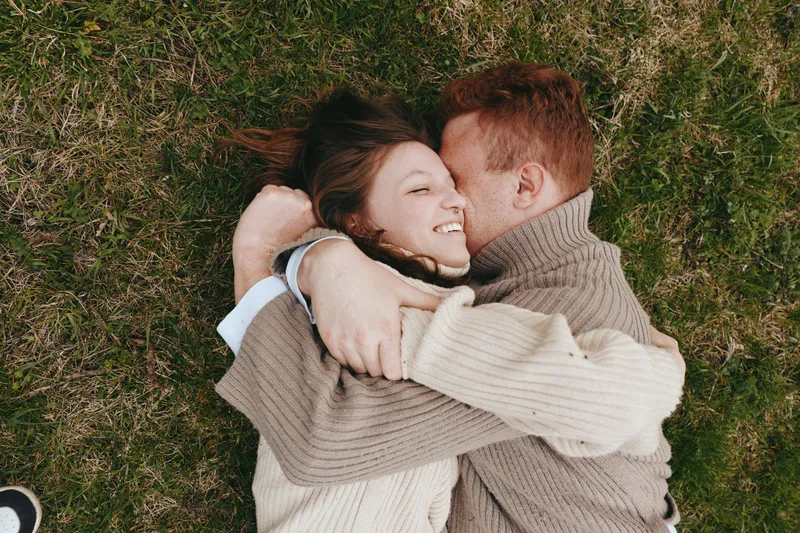 Smiling couple lying on grass embracing each other in cozy sweaters, enjoying outdoor relaxation and affection. Warm natural light and candid lifestyle moment convey romantic connection, casual