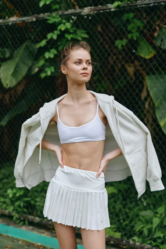 Young woman in sporty white outfit exuding confidence and joy, standing outdoors surrounded by lush green foliage, ideal for fitness and healthy lifestyle themes