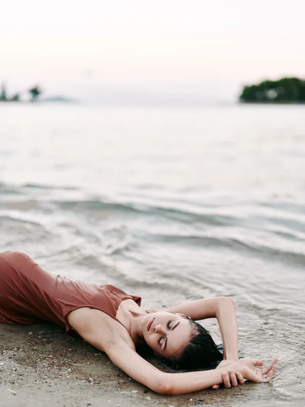 A serene beach scene shows a woman in a flowing dress lying on the sand, relaxed and contemplative as gentle waves roll in and a calm horizon stretches ahead.