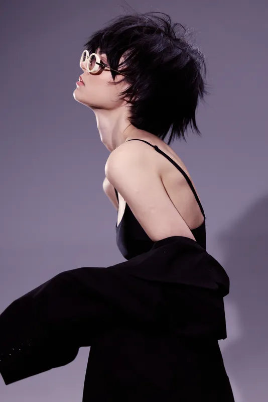Stylish woman with short hair wearing sunglasses and black outfit posing dramatically in studio lighting.