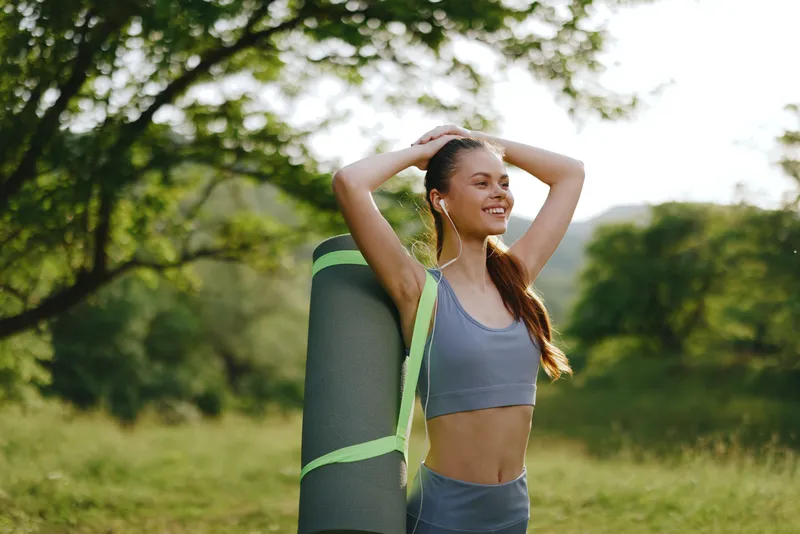 Smiling young woman with a yoga mat enjoying a sunny day outdoors, wearing sports attire and headphones, embodying wellness and fitness lifestyle