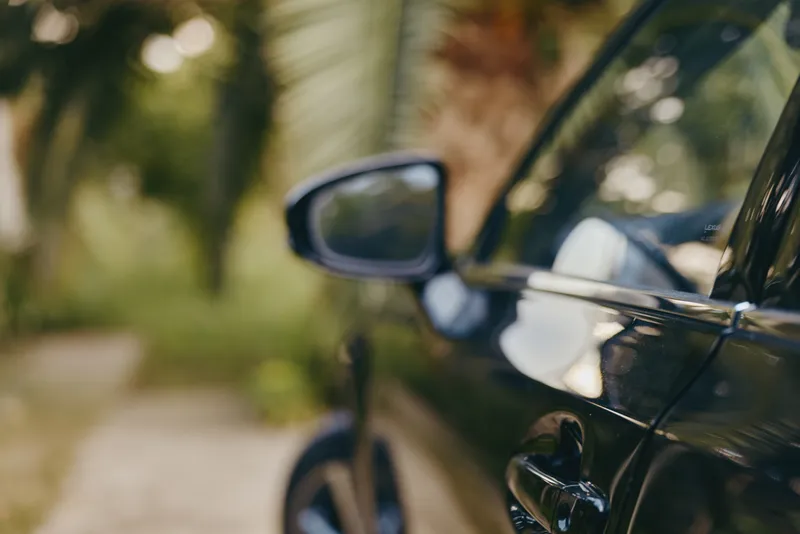 Black car side mirror and door handle closeup with glossy reflection and blurred palm bokeh, tropical outdoor setting. Modern compact sedan detail for travel lifestyle imagery.
