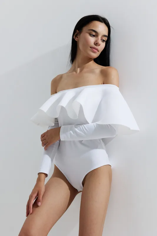 Woman sport fitness wearing white off-shoulder bodysuit, slender and poised, posing against a minimalistic light background with soft natural lighting.
