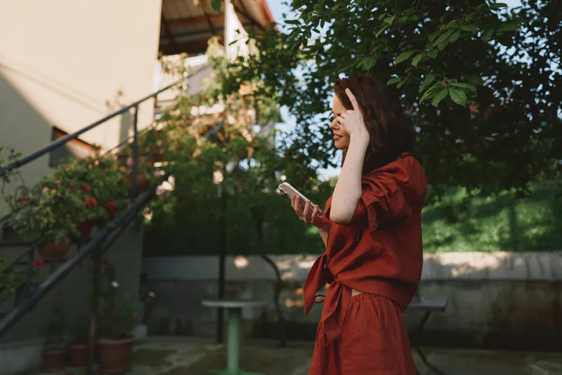 Cheerful woman outdoors, smiling while checking her phone, warm sunlight, relaxed posture, red dress, lush garden background, natural colors, casual lifestyle moment