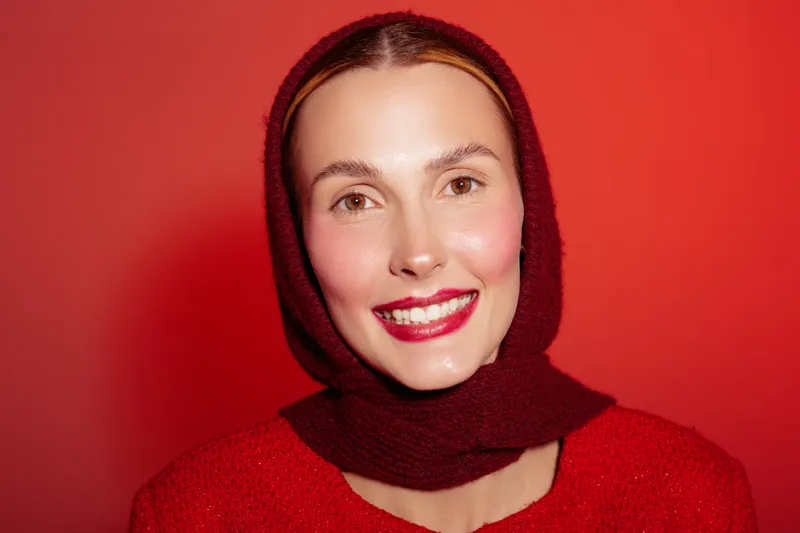 Smiling woman with red scarf and sweater poses against red background. Close-up portrait with natural makeup and bright lipstick. Concept of happiness, fashion, and winter style.