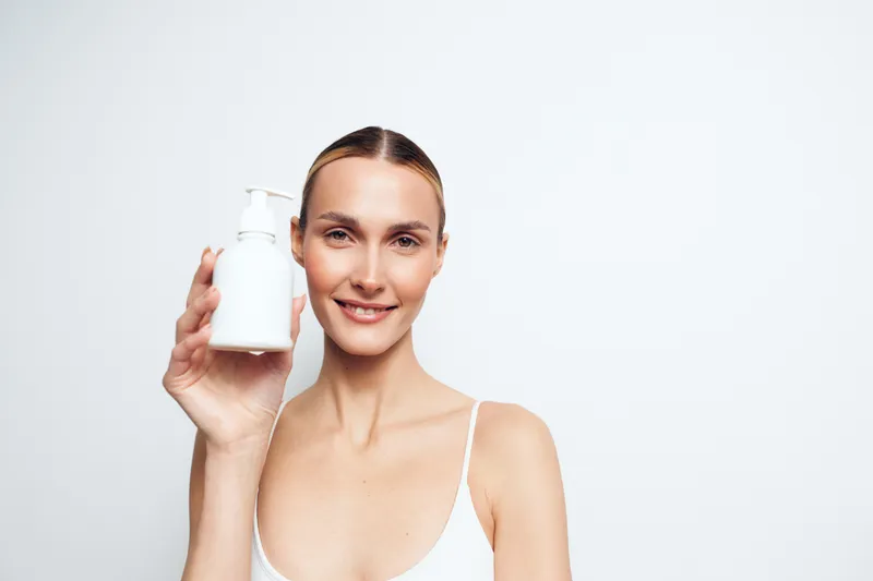 Young woman with brunette hair smiles gently while holding a white lotion bottle against a plain white background in a minimal skincare concept.