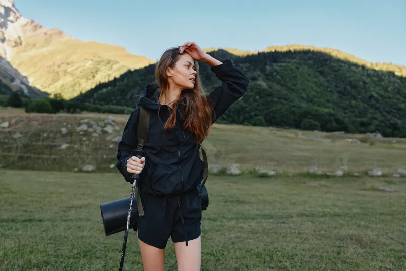 A young woman stands in a mountain meadow, wearing a dark jacket and shorts, with a backpack and trekking pole, looking into the distance and enjoying the outdoors.