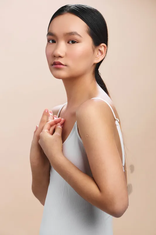 asian woman faces the camera in a studio portrait with warm neutral background. she wears a light sleeveless top, exuding calm confidence and a soft, inviting gaze.