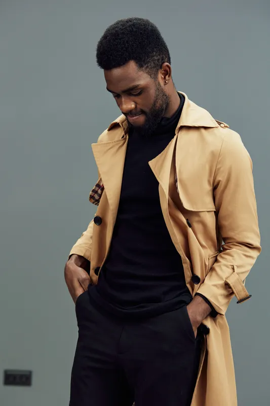 Stylish African man in a beige trench coat and black turtleneck, displaying a thoughtful expression against a muted green background