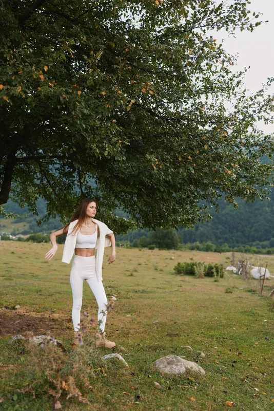 Young woman in white denim stands barefoot on a sunlit meadow beneath a large tree, enjoying a carefree outdoor moment and a peaceful, hopeful mood.