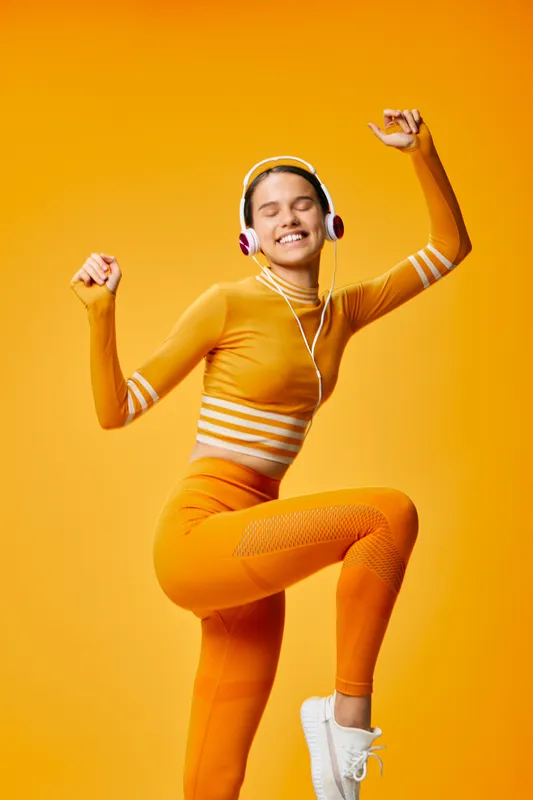 Energetic woman in bright orange athletic wear dancing with headphones against a vivid orange backdrop, showcasing fitness, movement and upbeat sport lifestyle.