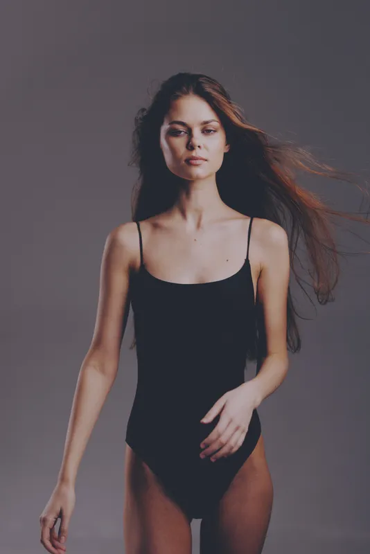 Young woman in black bodysuit posing confidently with natural makeup and flowing hair against a muted background
